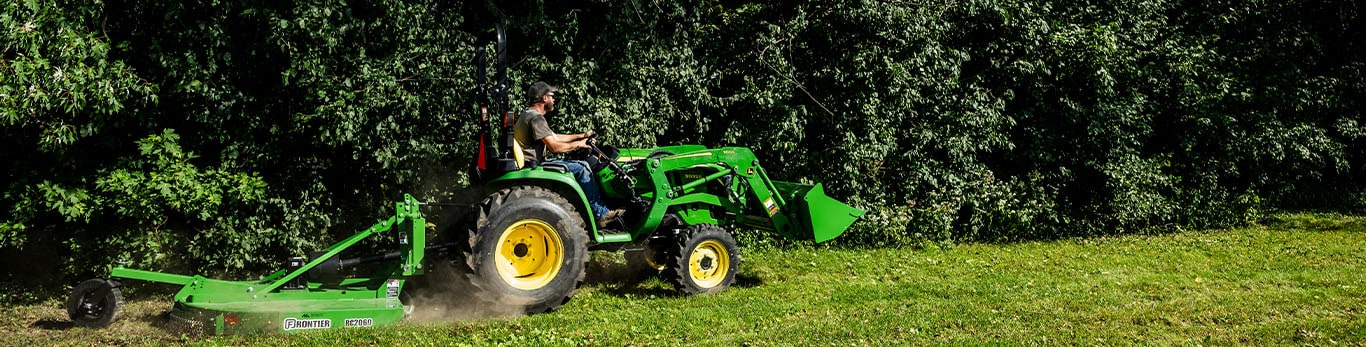 Une faucheuse rotative tract&eacute;e par un tracteur compact vert, coupant l'herbe le long d'une lisi&egrave;re bois&eacute;e.