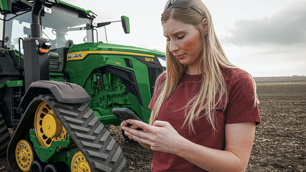 Une agricultrice se tient à côté d’un tracteur à chenilles John Deere 8RX 410 et connecte son téléphone à son tracteur compatible Autonomy Ready