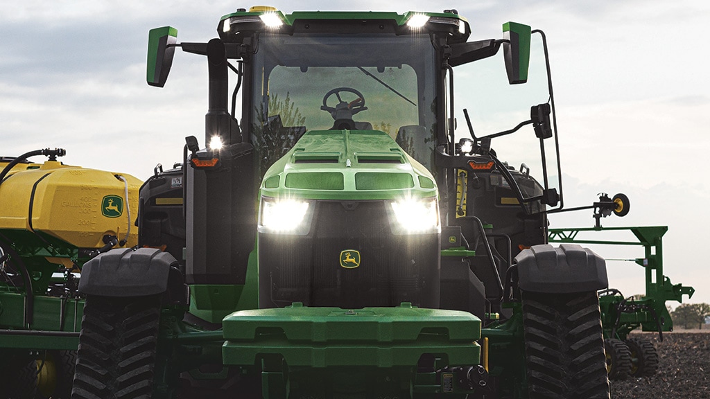 Tracteur 8RX haute puissance avançant phares allumés avec un planteur derrière.