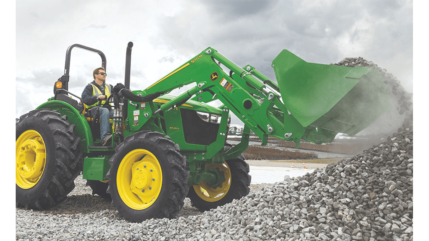 Une personne portant un gilet de sécurité utilise un chargeur frontal fixé à un tracteur vert pour prélever des cailloux dans un tas.
