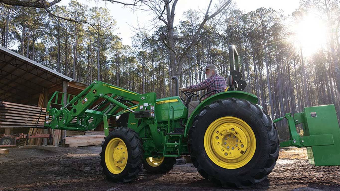 Un tracteur vert utilise un lève-palettes pour déplacer des planches dans une zone de stockage ouverte.
