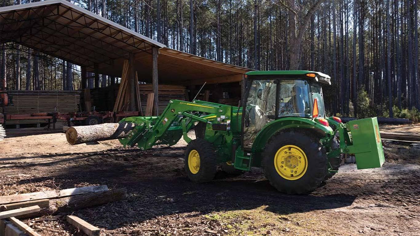 Un tracteur vert utilise un grappin pour déplacer des rondins de bois dans une zone de stockage ouverte.