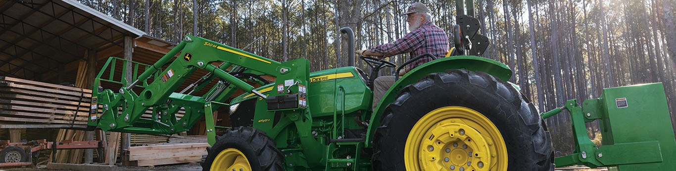 Un tracteur vert utilise une fourche à palette pour déplacer des planches sous une zone de stockage ouverte.