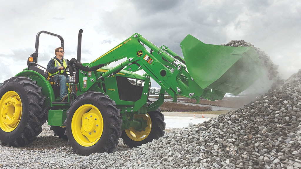 Une personne portant un gilet de sécurité utilise un chargeur frontal fixé à un tracteur vert pour prélever des cailloux dans un tas.