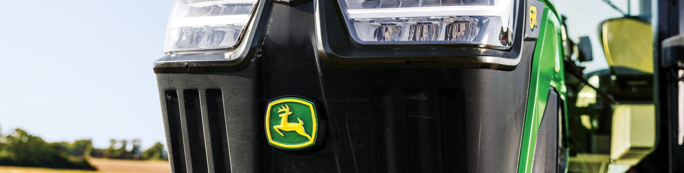 Front grille of John Deere tractor with logo, headlights, and field landscape in background.