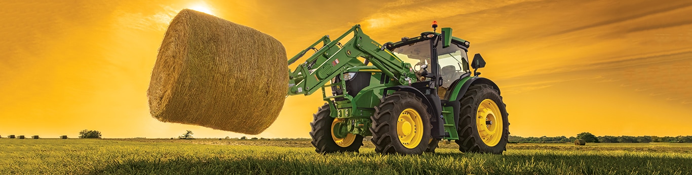 Tracteur 6M 125 avec chargeur soulevant une balle de foin au coucher du soleil