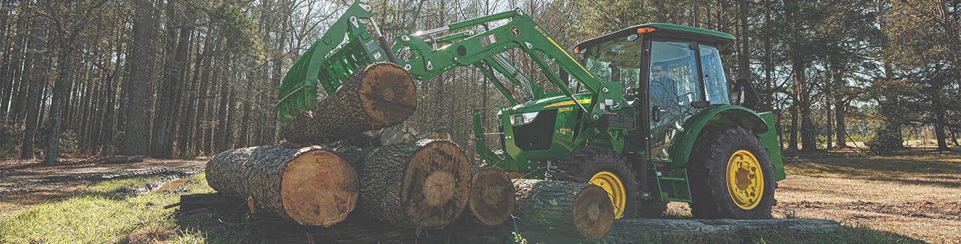 Un tracteur vert équipé d'une pince avant lève de grosses bûches dans une zone boisée.