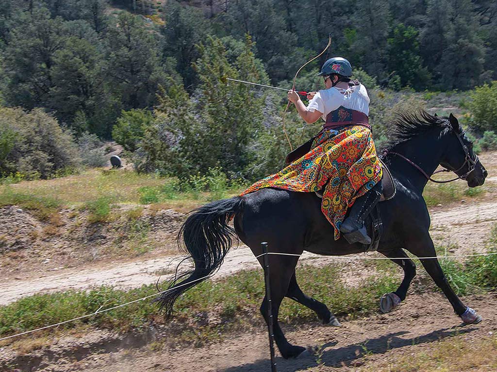 archer on horseback preparing a shot while horse is galloping with greenery in the background