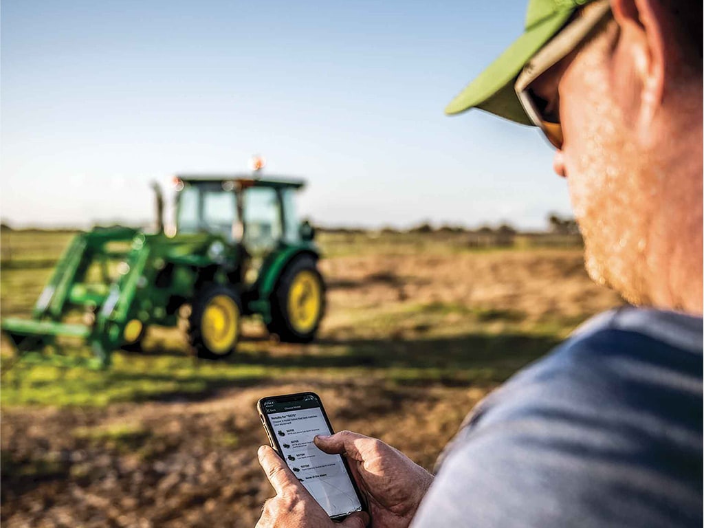 Homme dans un champ utilisant l’application John Deere Property Center avec un tracteur en arrière-plan