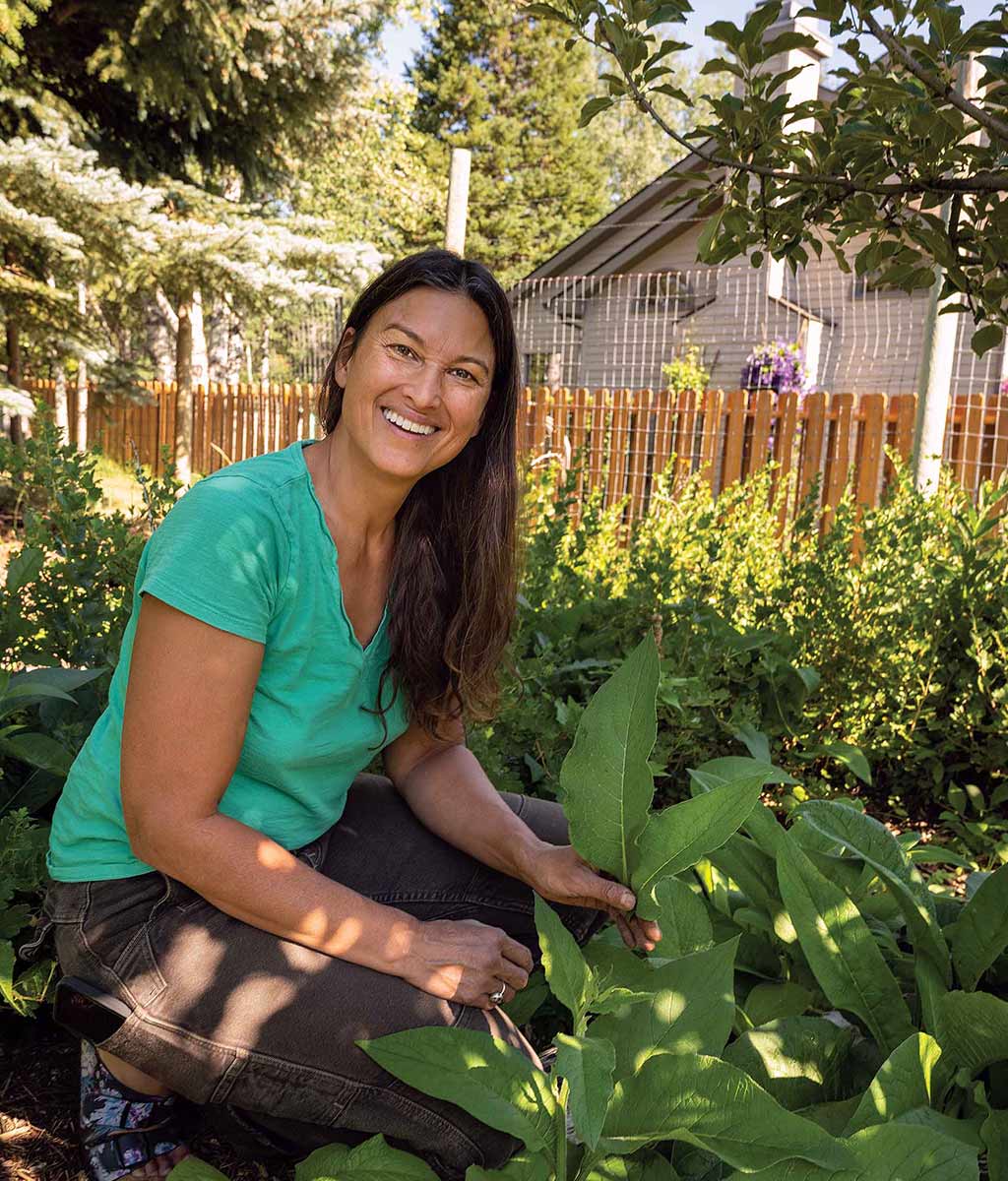 woman with long dark hair squatting and smiling holding large plant leaves