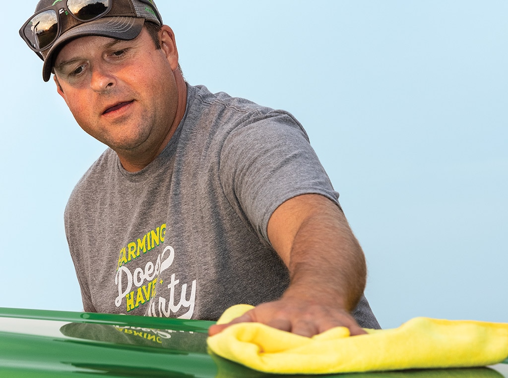 man with baseball hat polishing green Deere equipment with yellow towel