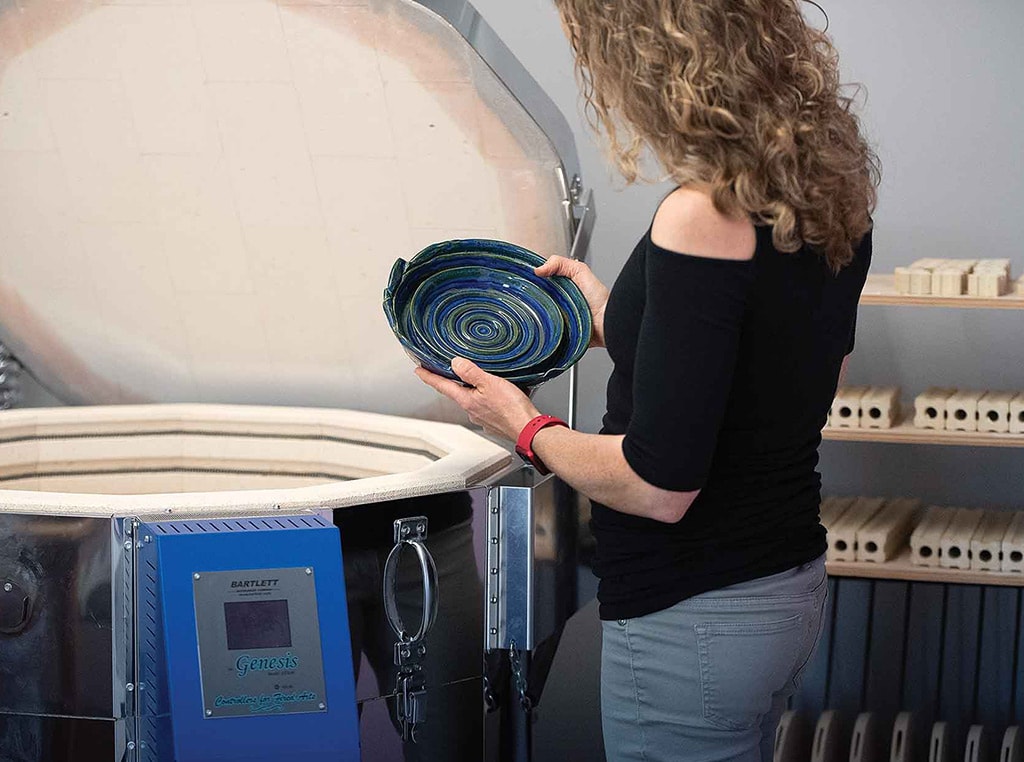 woman standing in front of open kiln inspecting blue clay bowl