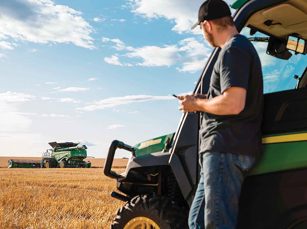 Homme debout tenant un téléphone cellulaire, à côté d’un véhicule utilitaire Gator de John Deere, qui regarde une moissonneuse-batteuse dans un champ