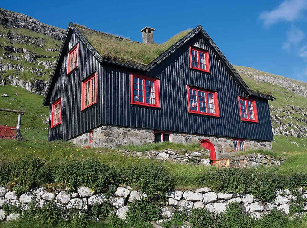 dark blue house with red window frames on grassy hillside with stone wall in front