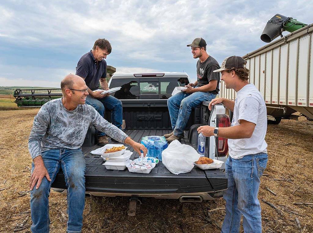 four men standing and sitting around truck bed eating out of styrofoam boxes