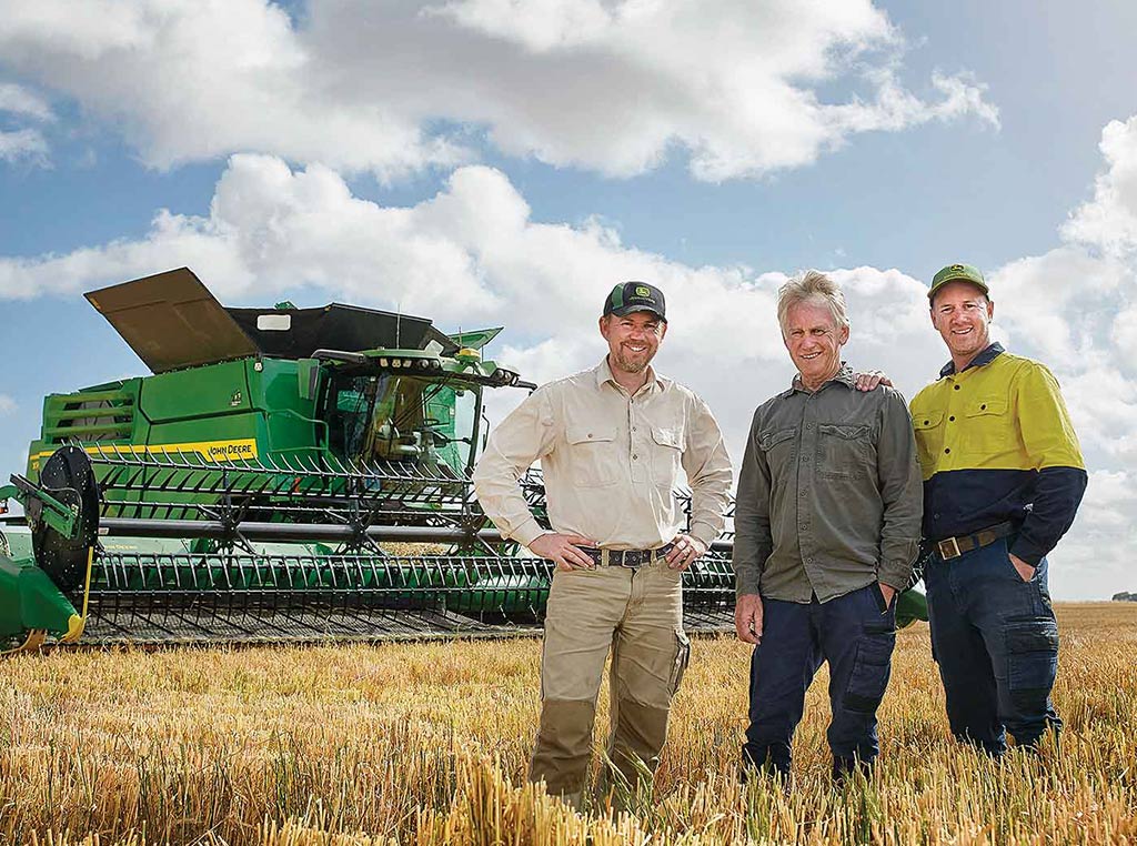 Trois hommes debout dans un champ, devant une moissonneuse-batteuse de John Deere