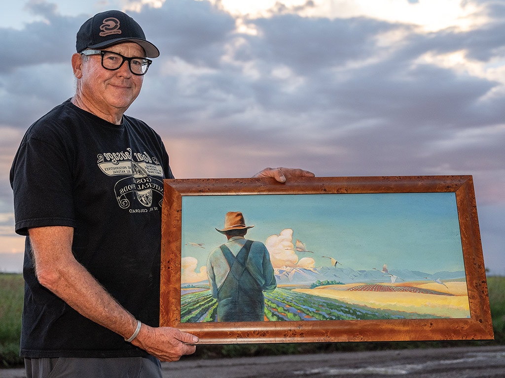 Person with black tshirt, baseball cap, and glasses presenting painting of farmer facing field of crops from behind