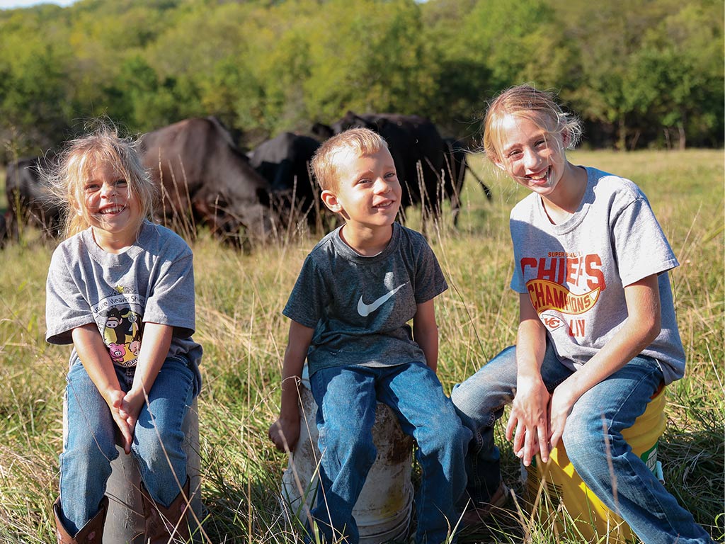 Trois enfants souriants assis sur des seaux dans un champ avec du bétail