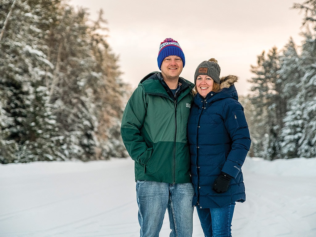 Un couple en vêtements d’hiver posant dans la neige entre deux rangées de conifères