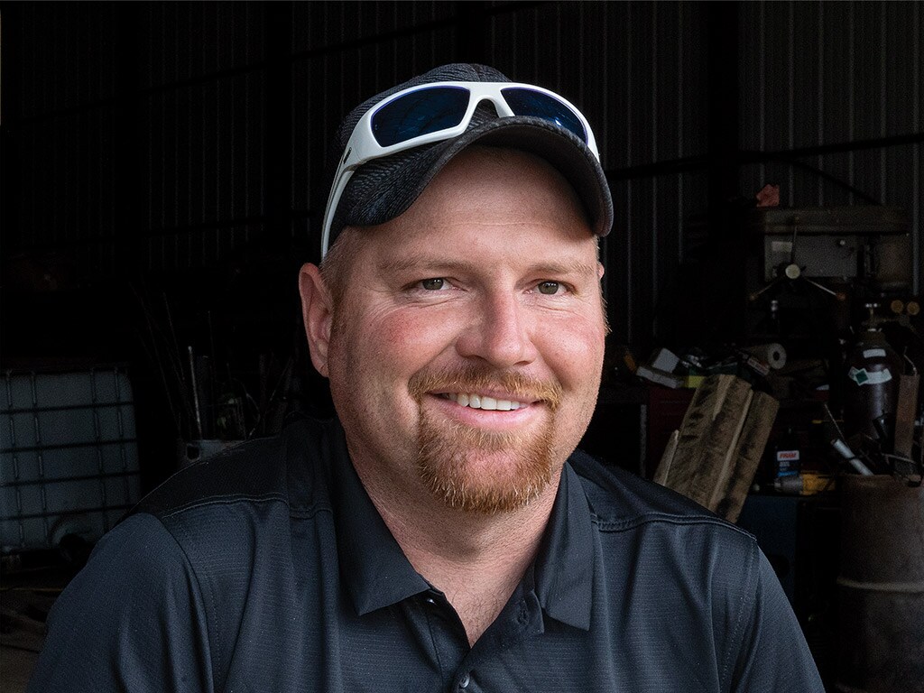 Person smiling with red goatee, baseball cap and white sunglasses atop their head