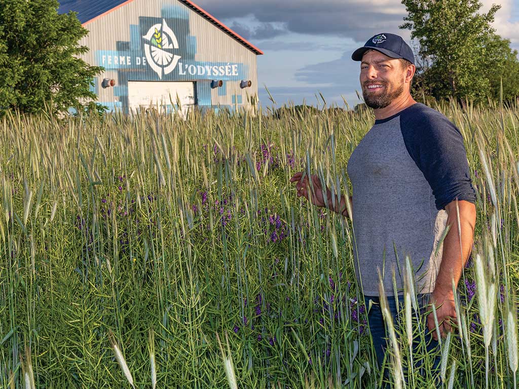 Barbu souriant portant une casquette de baseball dans un champ d’herbes hautes avec des arbres et un entrepôt en arrière-plan