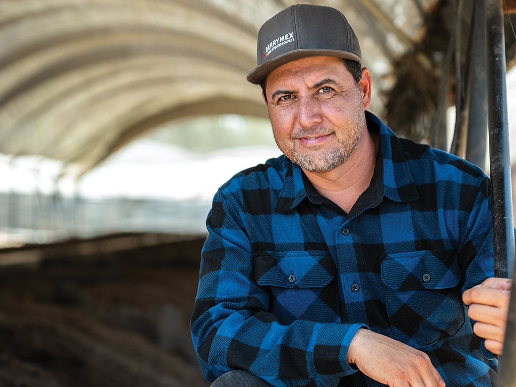 Person smiling with baseball cap and black and blue plaid flannel shirt crouched in compost tent