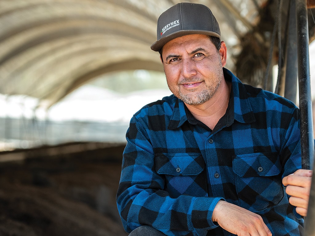 Personne souriant avec une casquette de baseball et une chemise en flanelle à carreaux noire et bleue accroupie dans une tente de compost