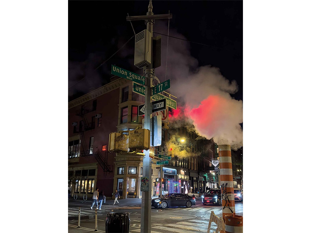 a city intersection at night with steam rising from an orange and white pipe