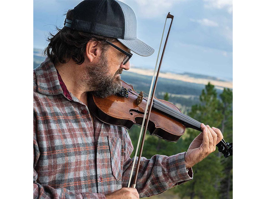 person in a baseball cap and glasses playing the violin with trees and hills in the distant background