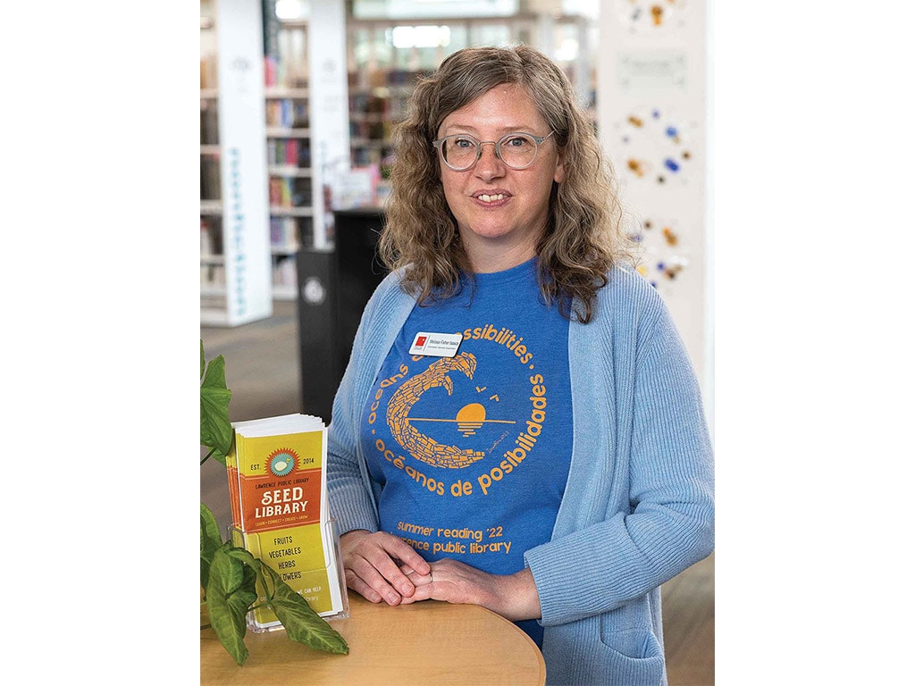 smiling person with glasses standing next to a stack of pamphlets for the seed library