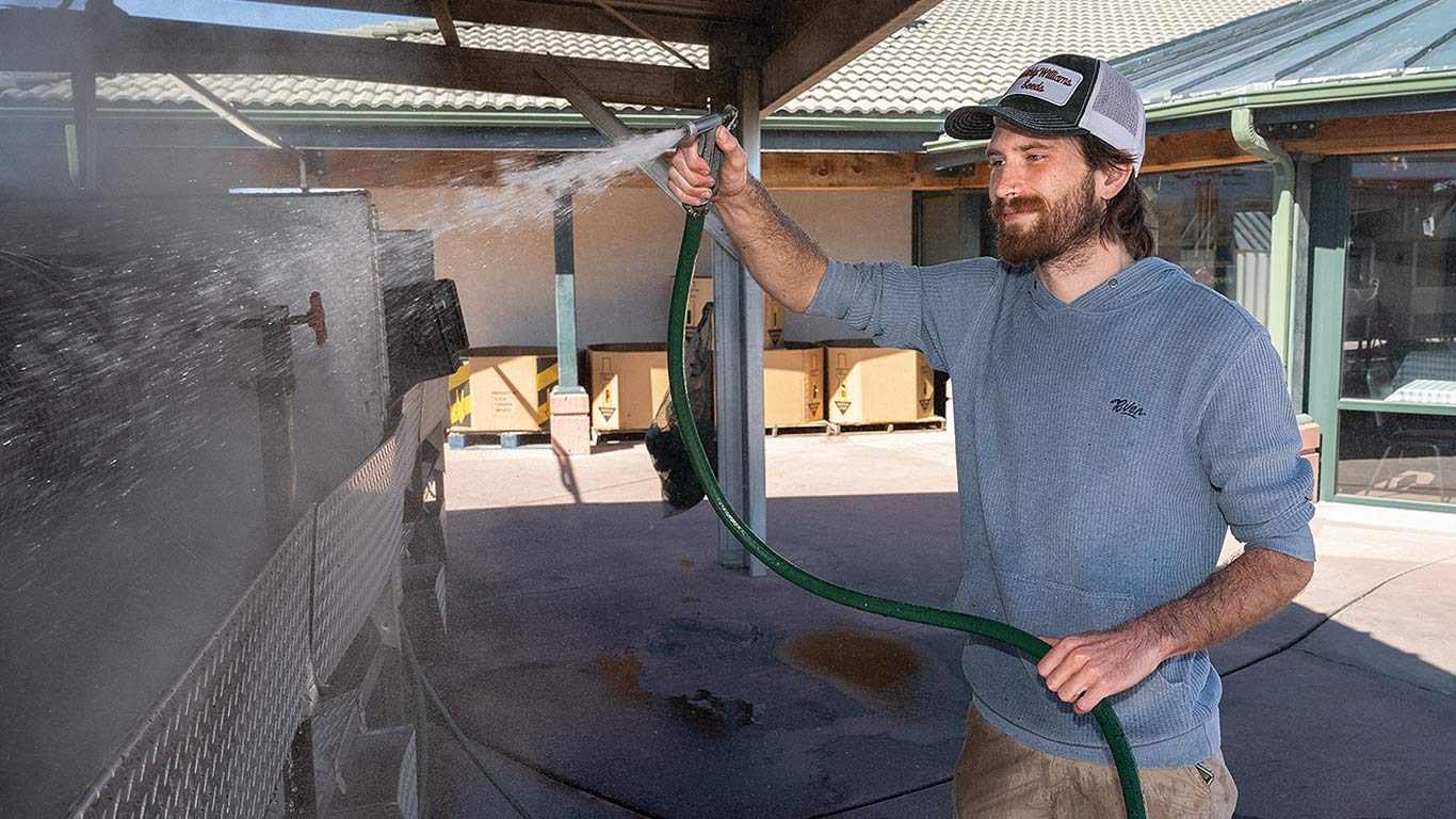 Person in blue sweater and cap sprays water with hose on outdoor surface.