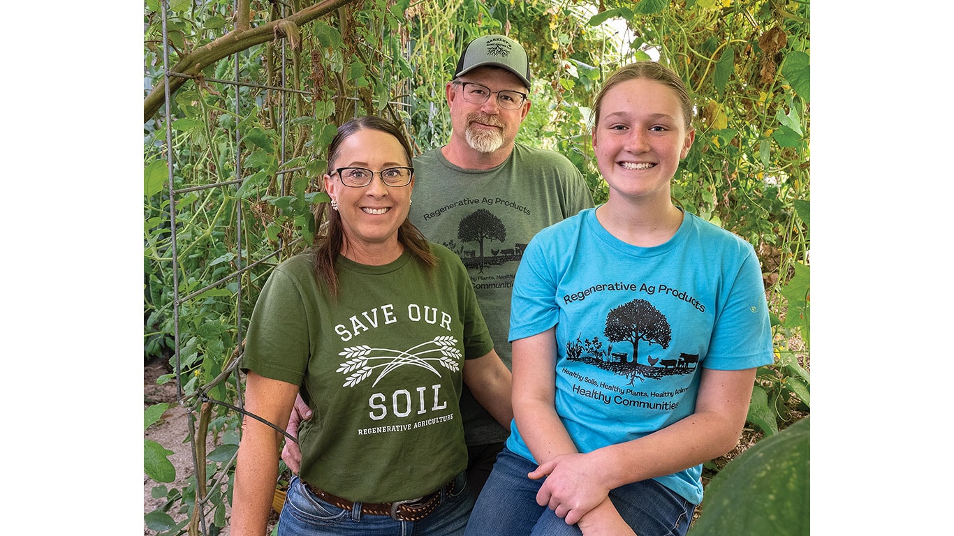 Three individuals stand together in a lush garden, wearing t-shirts promoting agriculture practices.
