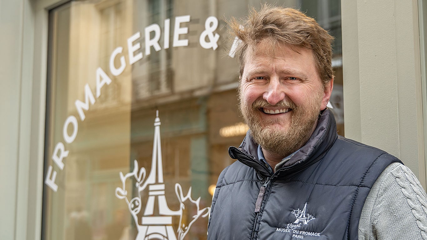 Une personne souriante devant la vitrine d'une boutique « FROMAGERIE ».