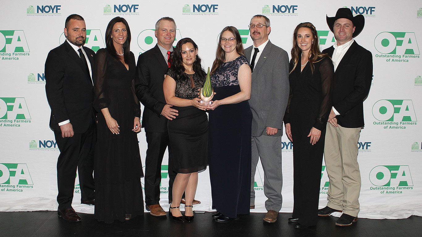 Huit personnes posent lors d’un événement en tenant un prix; le logo de l’OFA en arrière-plan.