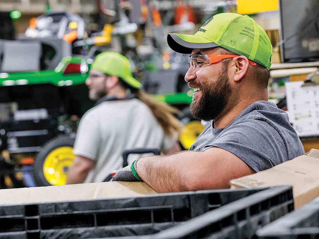A smiling John Deere worker with machinery in the background