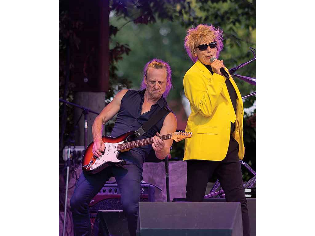 A singer in a bright yellow jacket performs on stage beside a guitarist with an electric guitar.