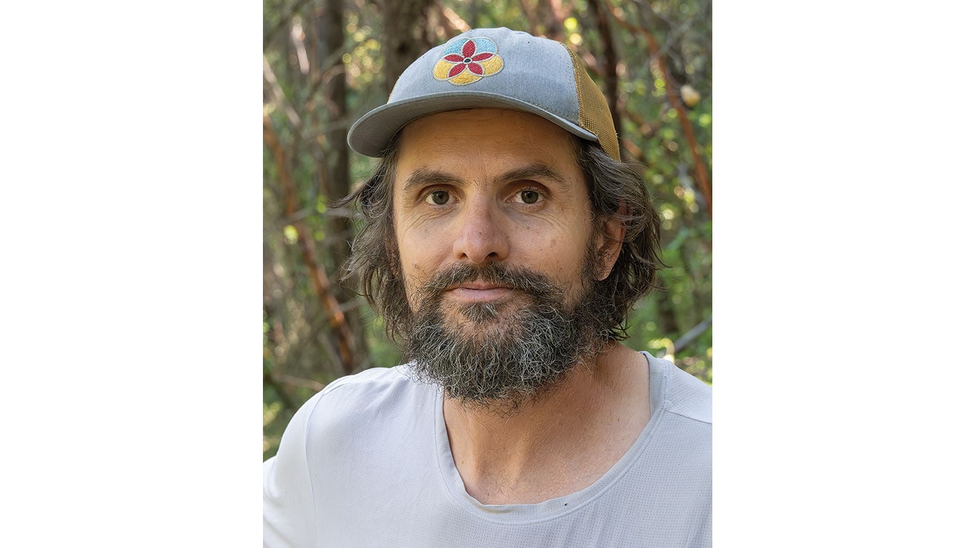 Bearded person wearing a cap, photographed outdoors with trees in the background.