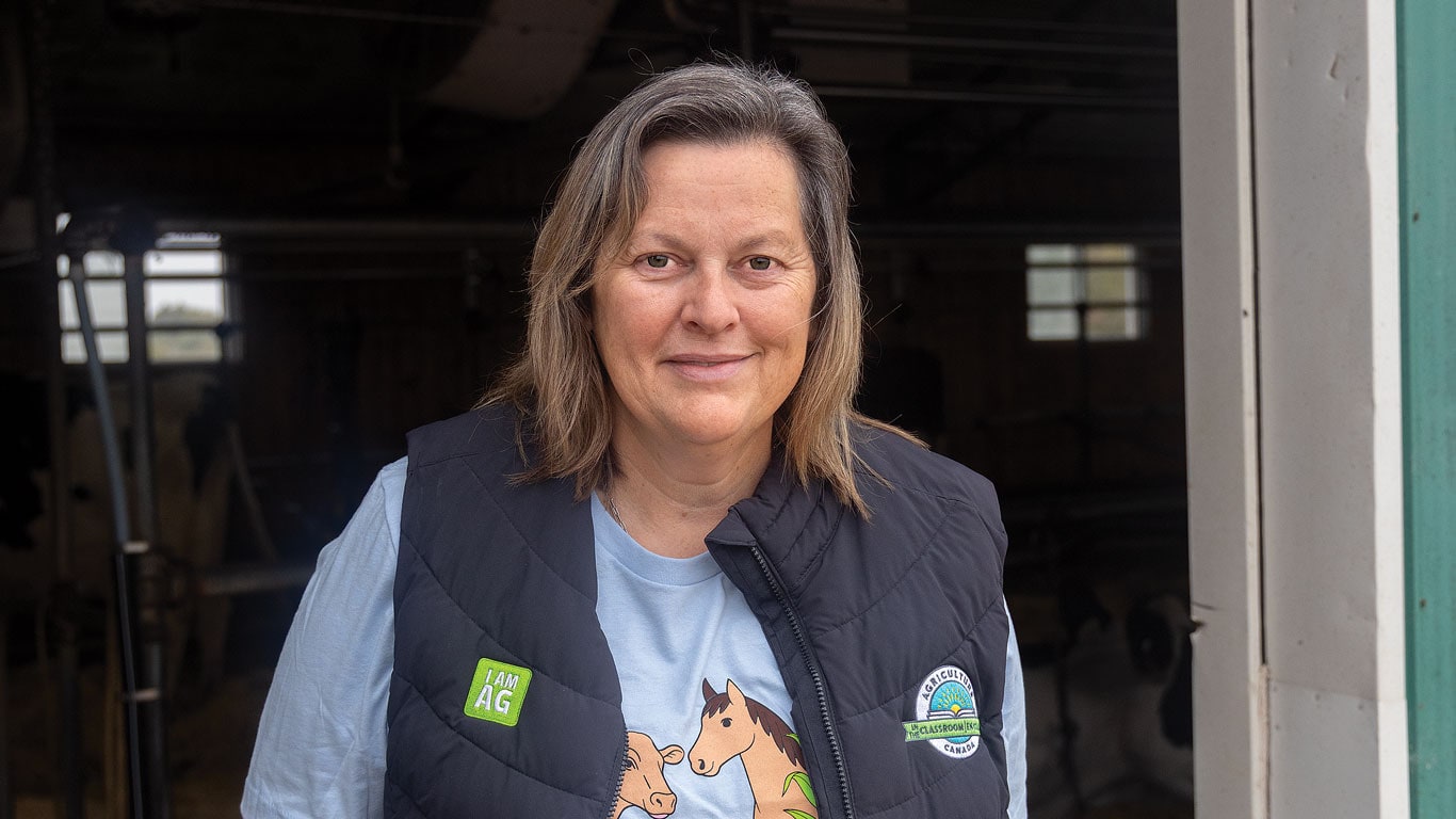 Woman wearing a vest stands in a barn doorway, smiling at the camera.