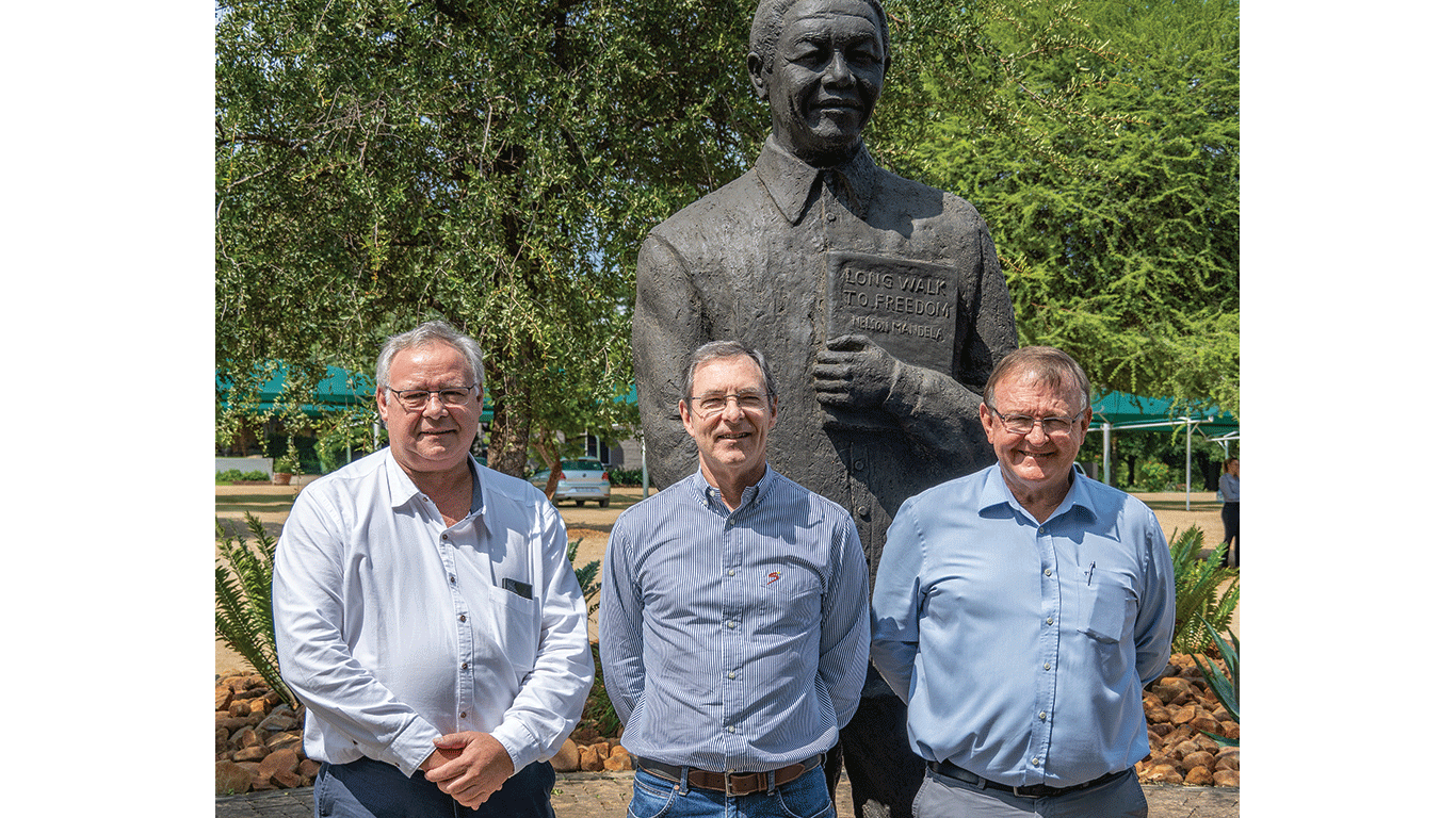 Trois personnes se tiennent devant une grande statue d'un homme tenant un livre, en plein air, au milieu d'arbres et de plantes.