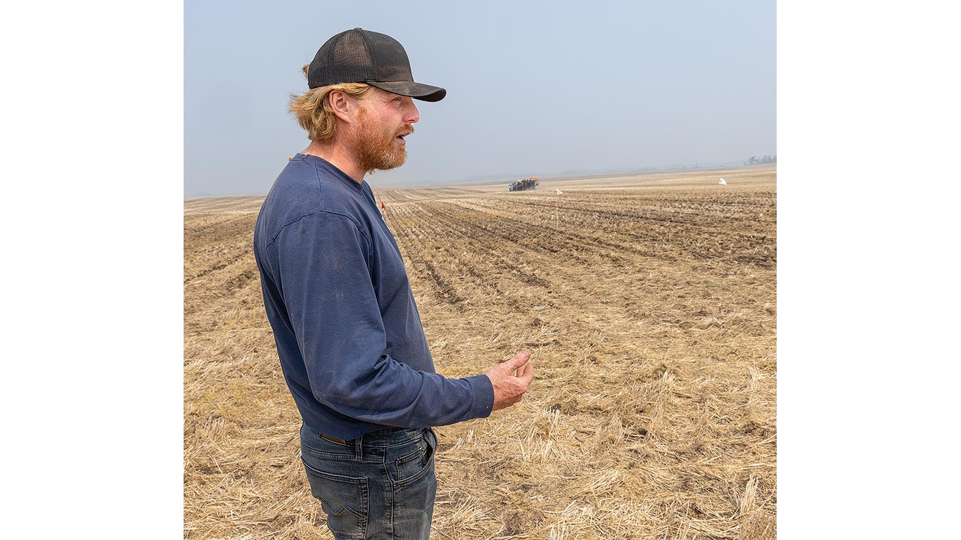 Un fermier debout dans un champ r&eacute;colt&eacute; examine la terre. Au loin, de la machinerie est au travail.
