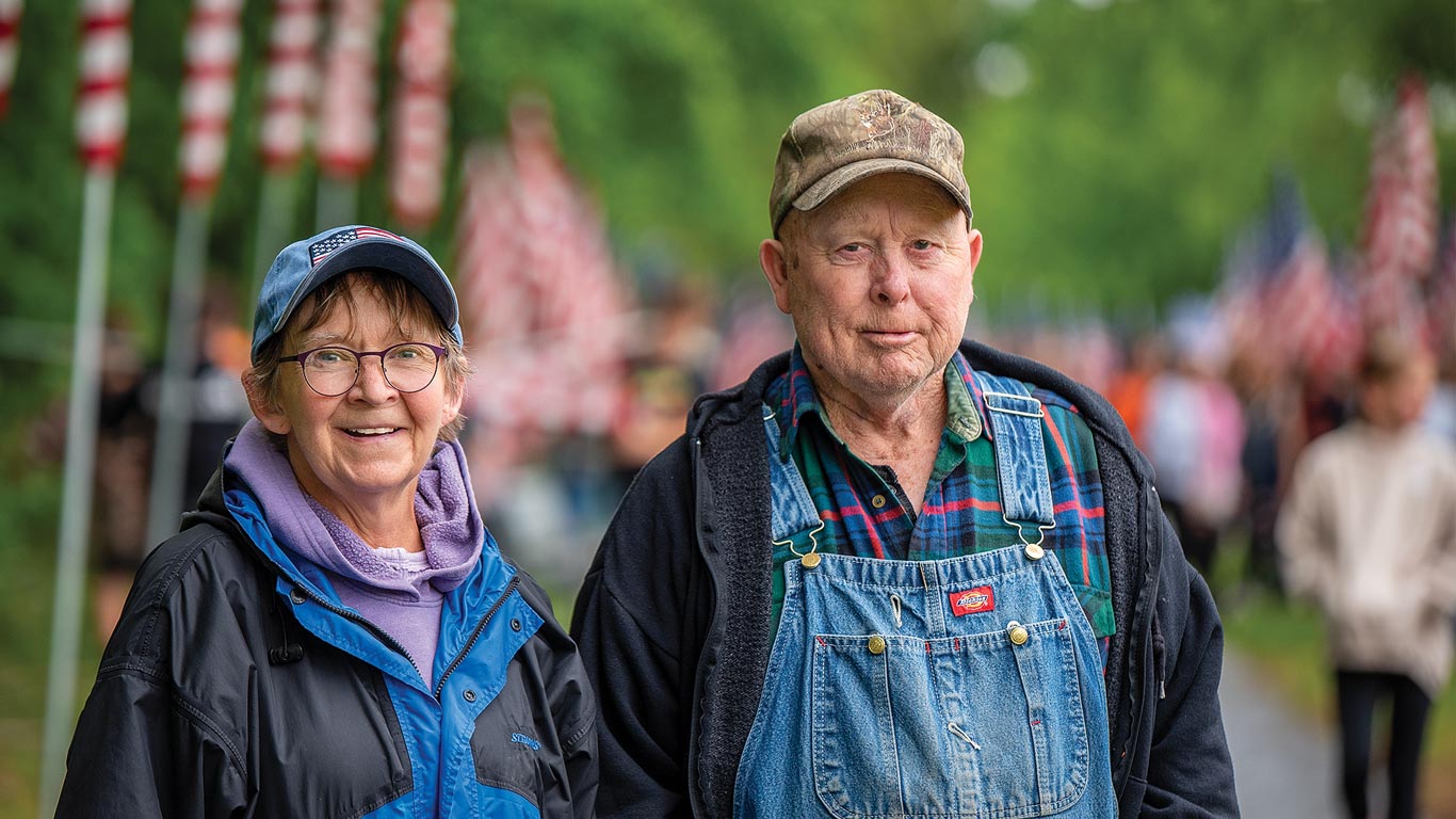 Older couple stands together along a flag‑lined walkway.