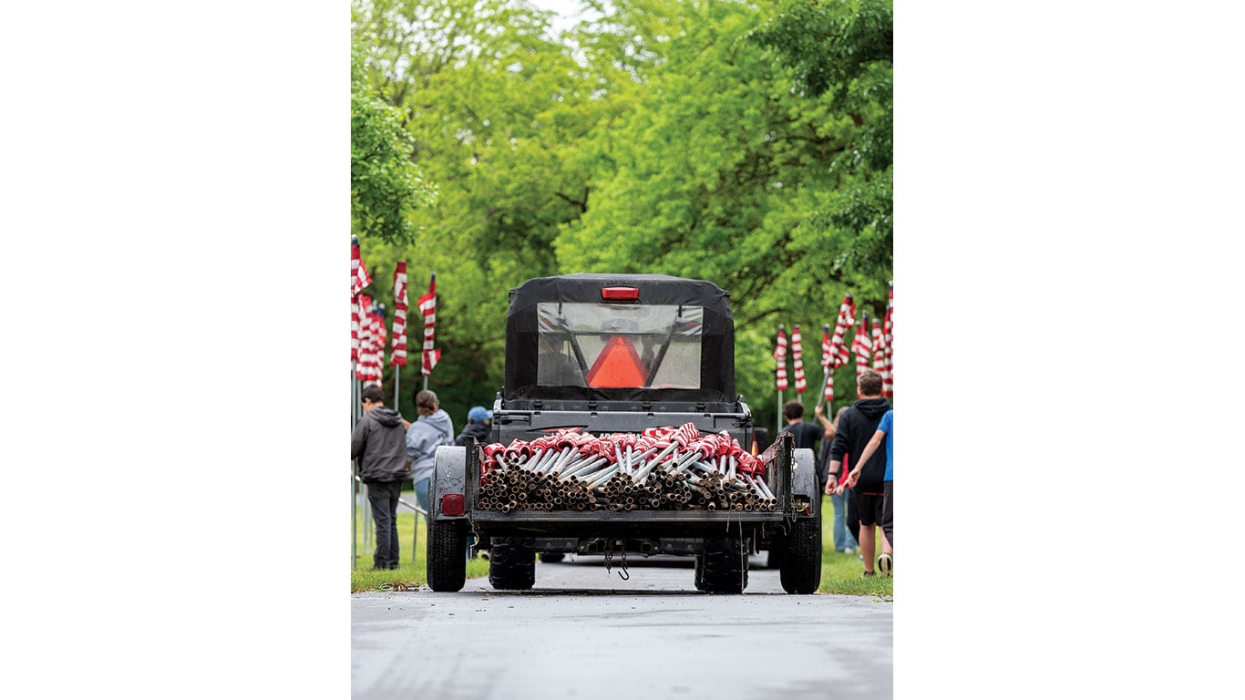 Vehicle carries rolled American flags along a tree‑lined path.