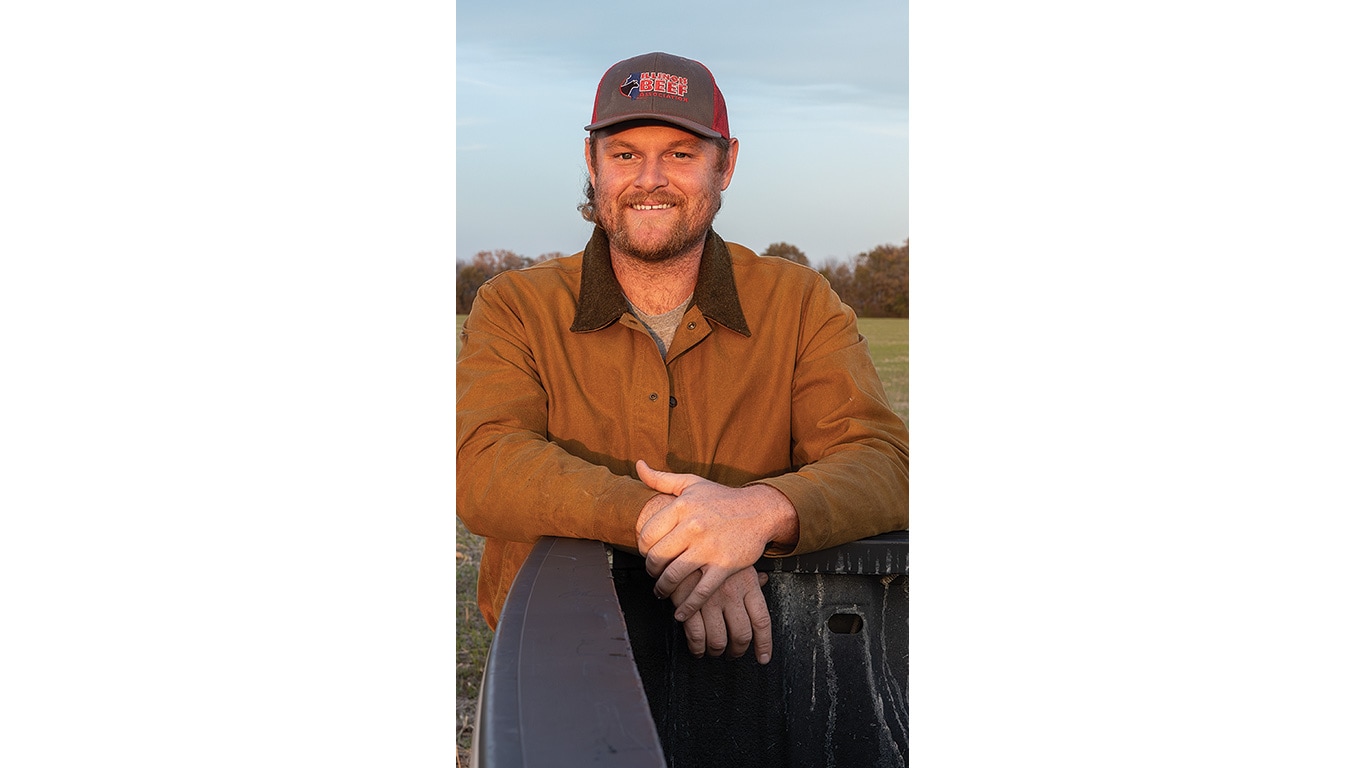 Farmer wearing a cap and jacket smiles while leaning on a truck in an open field.