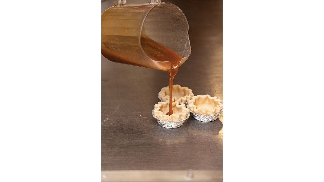Caramel filling being poured into miniature pie crusts on a stainless steel worktable.