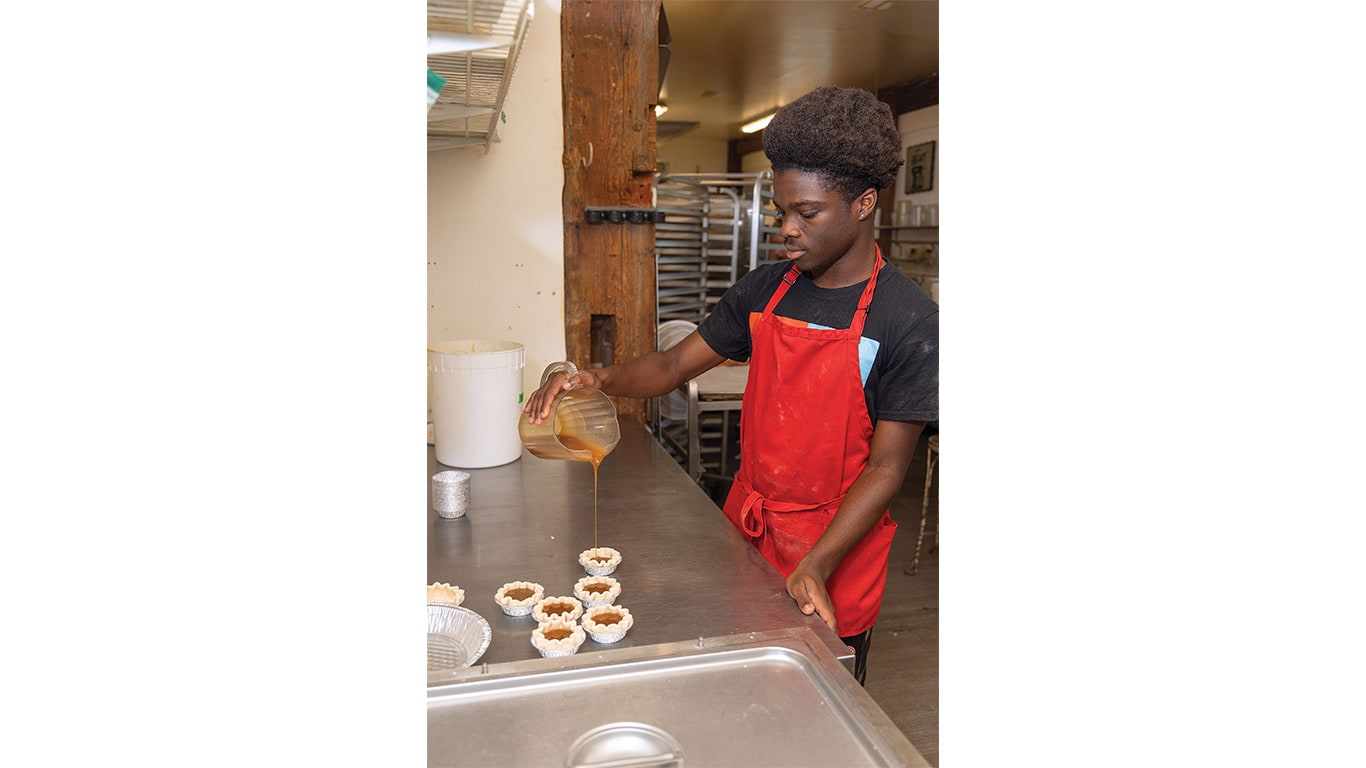 Person in a kitchen pouring filling into pie crusts on a stainless steel preparation table.