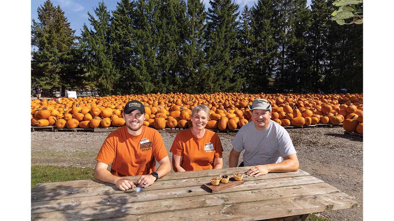 Three people seated at a picnic table with miniature pies in front of a large pumpkin display.