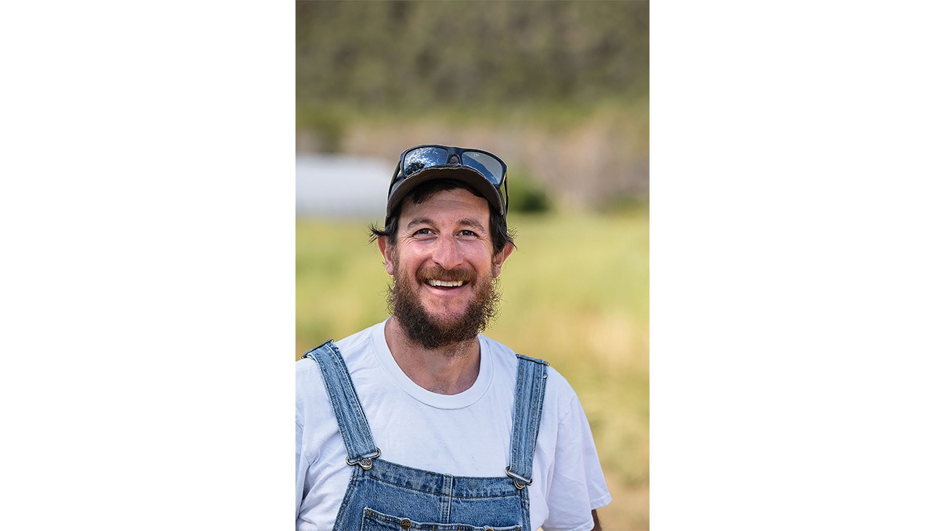 Person wearing overalls and a cap standing outdoors with a grassy field in the background.