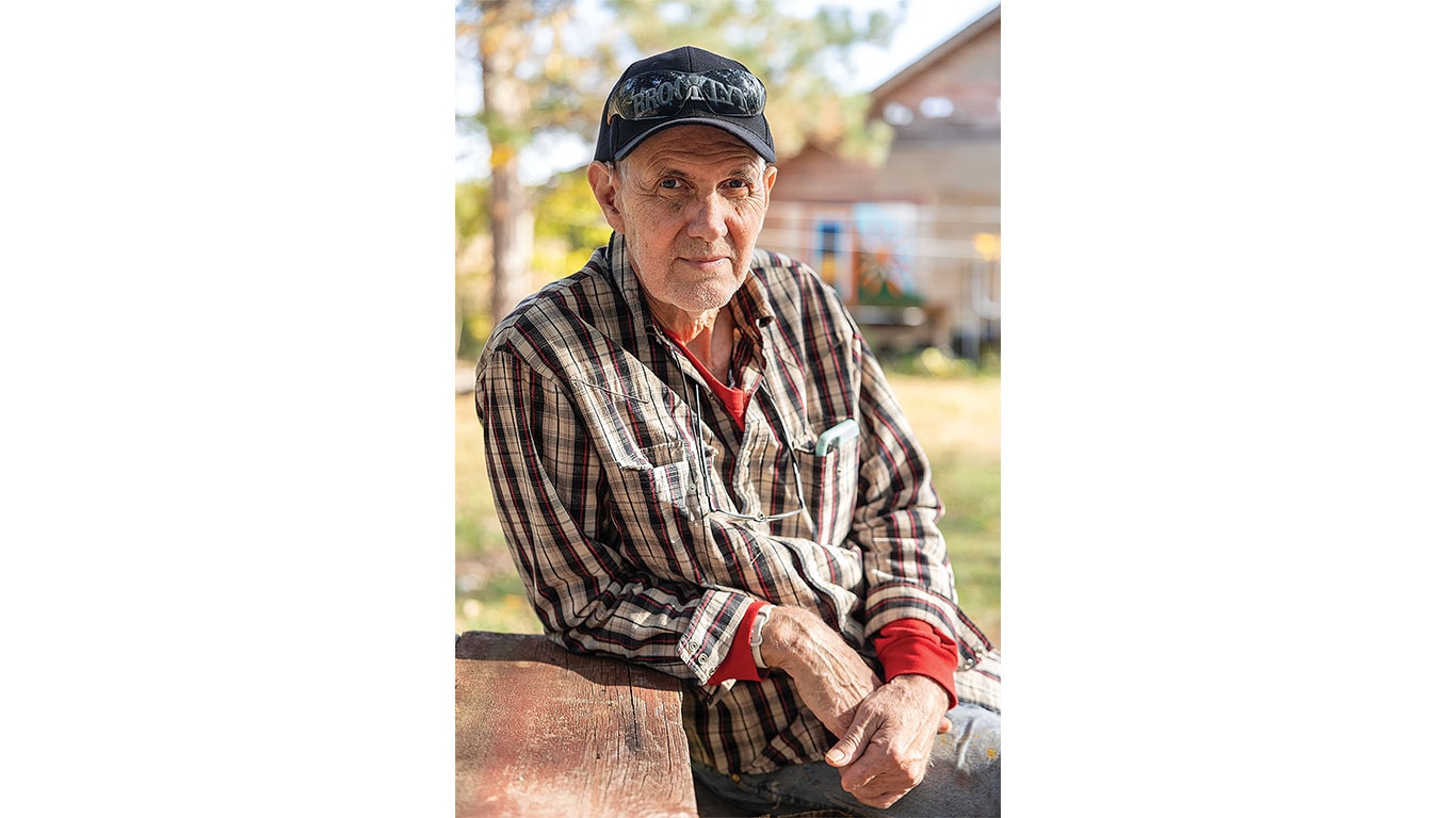 Person in a plaid shirt and cap seated outdoors beside a wooden bench.