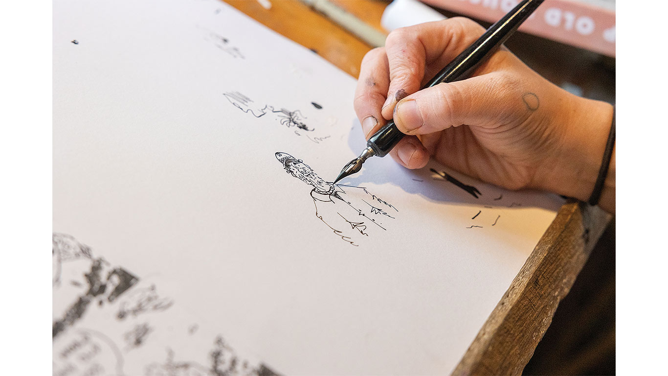 Close-up of a hand using a pen to draw illustrations on paper at a worktable.