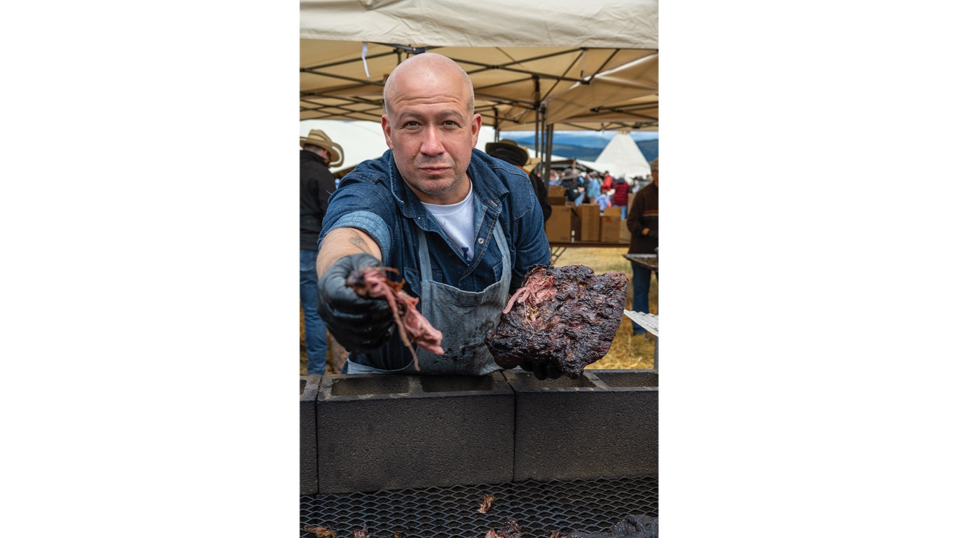 Person serving smoked meat at an outdoor cooking station beneath a canopy tent.